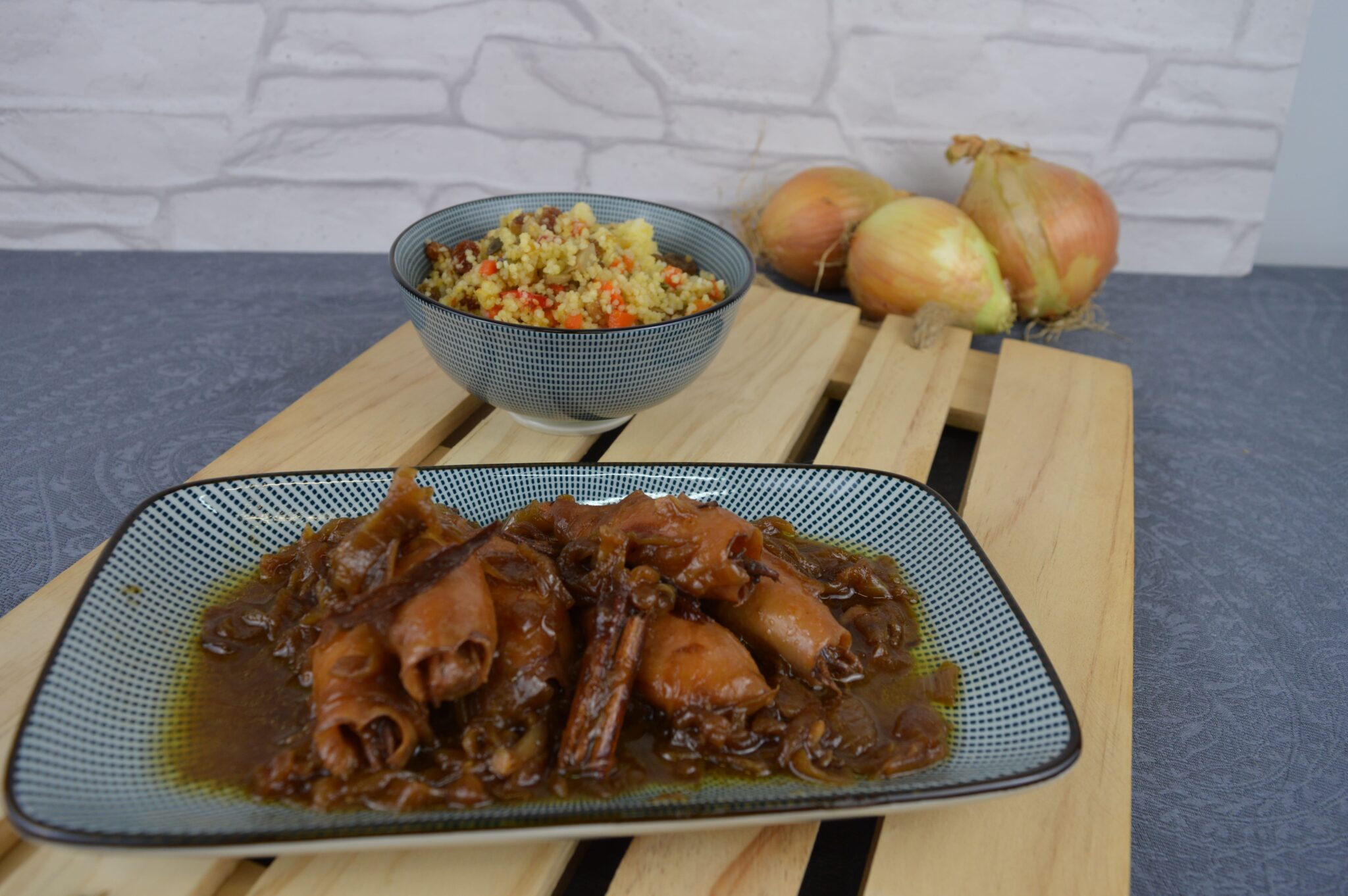 Chipirones encebollados a la canela en salsa oscura de cebolla con su rama de canela, servidos en plato alargado con un cuenco de cuscús de verduras y cebollas al fondo