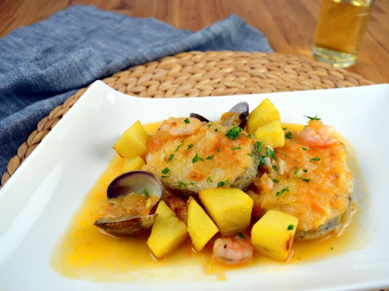Rodajas de merluza en salsa con almejas, gambas y dados de patata frita, servidas en plato blanco con perejil picado