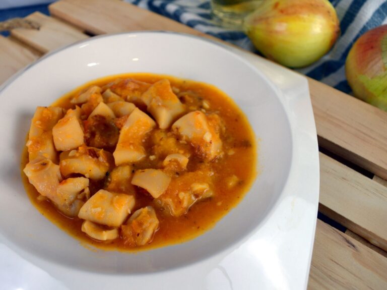 Sepia en salsa con majado de almendras servida en plato hondo blanco, con cebollas al fondo sobre tabla de madera