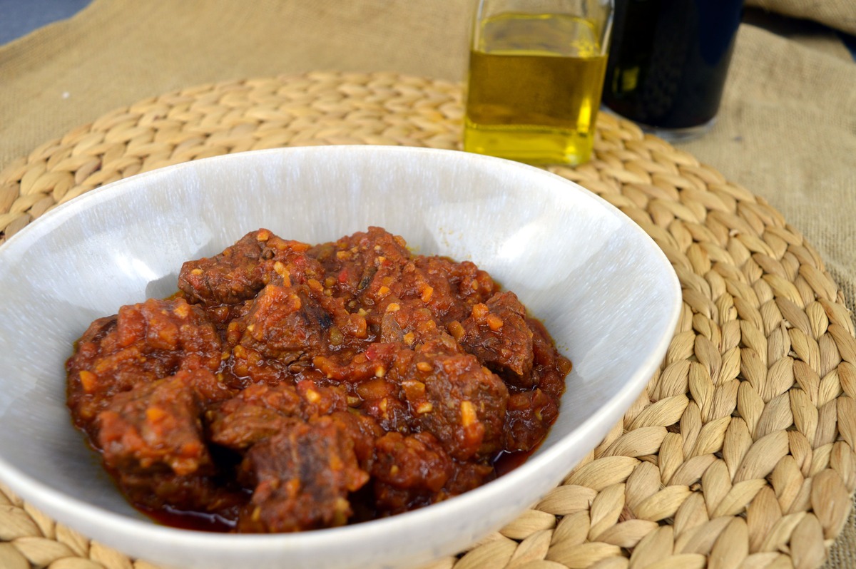 Ternera guisada en salsa de tomate y pimientos en cuenco de cerámica, con aceite de oliva y vino al fondo